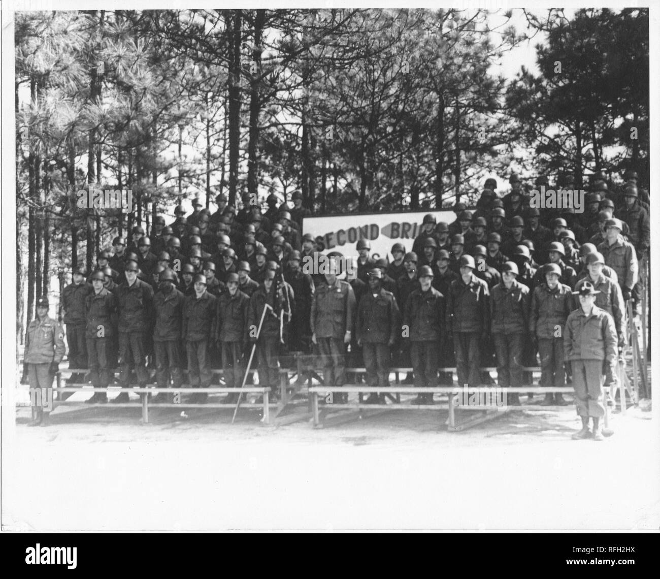 Fotografia in bianco e nero di un gruppo di militari Usa i soldati, indossano uniformi e caschi, insieme permanente sulle gradinate o gabbie, con alberi di pino visibile in background, oscurato un segno visibile al centro (probabilmente con il testo 'Seconda Brigata") e un soldato tenendo un contrassegno visibile al centro di primo piano, fotografato durante la Guerra del Vietnam, 1967. () Foto Stock