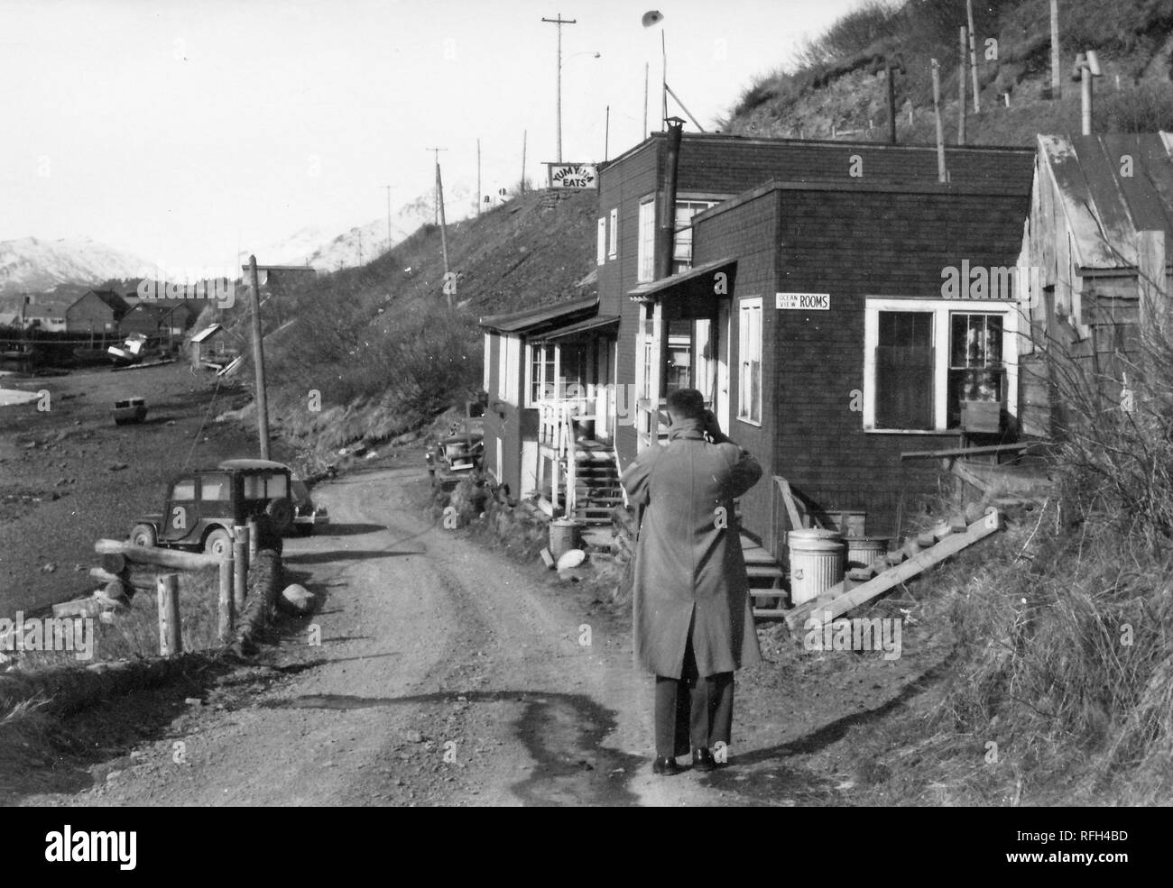 Fotografia in bianco e nero di un uomo, visto dal retro, indossando un cappotto lungo e in piedi sul bordo di una strada sterrata, con le braccia sollevate alla sua testa (probabile utilizzando una telecamera) con una linea costiera e la vecchia auto a sinistra, vari piccoli edifici in legno (con i segni " Camere " e " Yum Yum mangia') a destra e in più edifici in legno e montagne innevate visibile sullo sfondo, fotografato durante una battuta di caccia e viaggio di pesca si trova in Alaska, Stati Uniti d'America, 1955. () Foto Stock