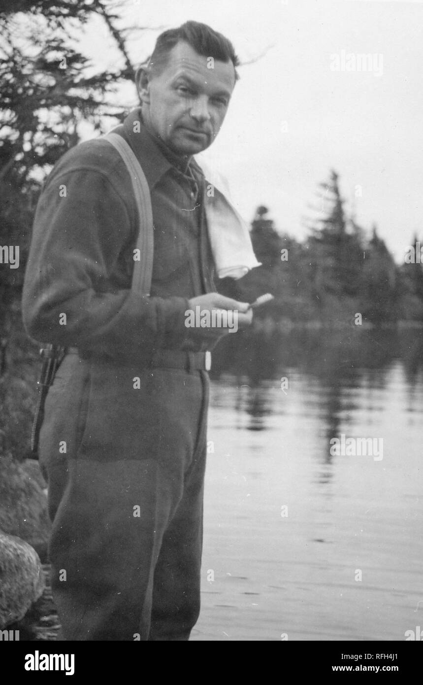 Fotografia in bianco e nero di un uomo di mezza età, sta in piedi in un litorale, in tre quarti di lunghezza vista di profilo, con la testa rivolta verso la telecamera, che indossa pantaloni con bretelle su una maglia di lana e tenendo uno spazzolino da denti in una sola mano, con alberi e fogliame visibile sullo sfondo, fotografato durante una battuta di caccia e viaggio di pesca si trova in Alaska, Stati Uniti d'America, 1955. () Foto Stock
