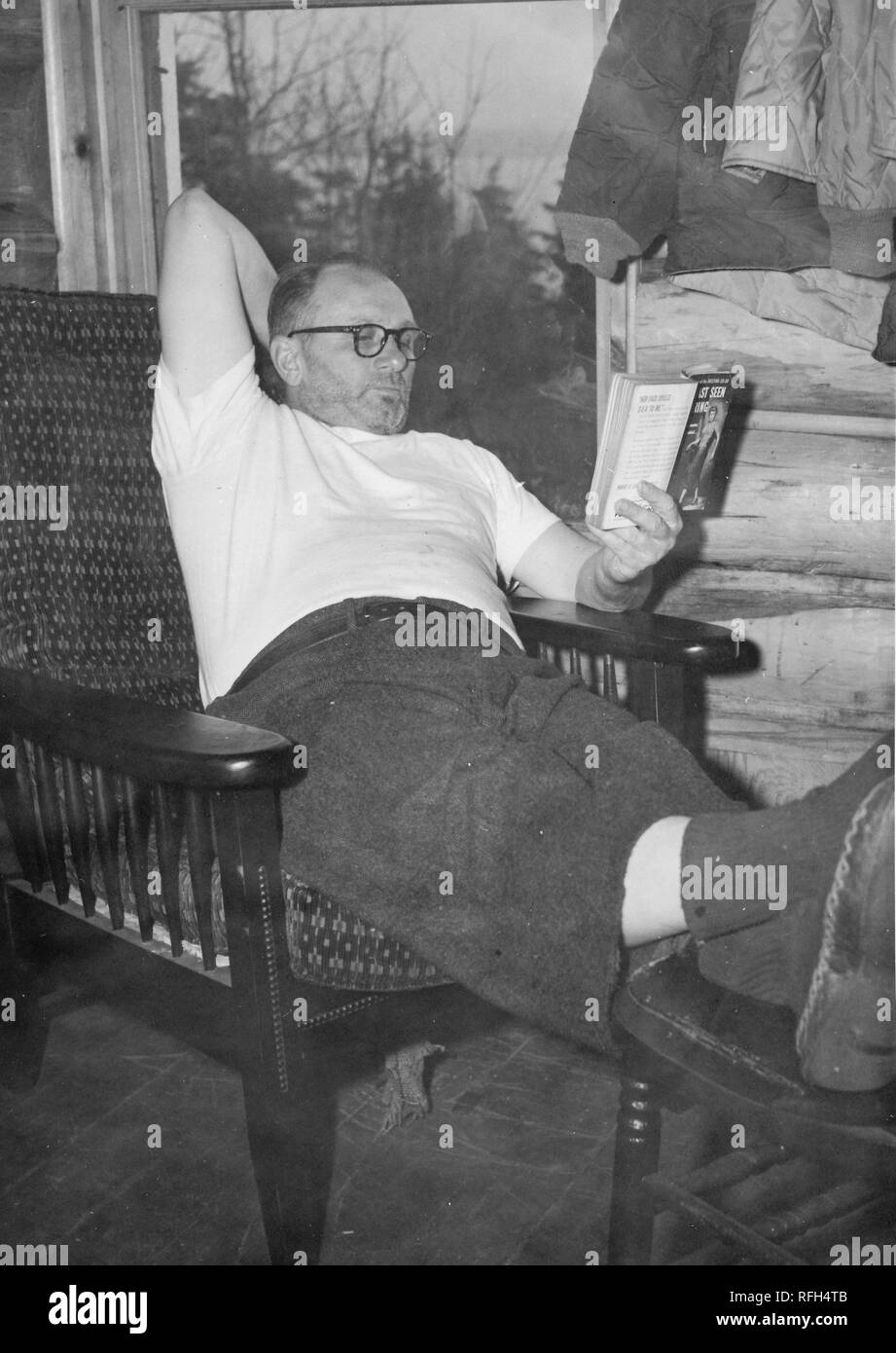 Fotografia in bianco e nero di barba lunga di un uomo di mezza età, in piena vista di lunghezza, indossando occhiali, una t-shirt bianco e pantaloni di lana, la lettura di un libro in brossura reclinabili mentre in un bosco una selleria sedia, con i suoi piedi appoggiato su uno sgabello; con log di mura e di una porta o una finestra attraverso la quale gli alberi di conifere sono visibili sullo sfondo, fotografato durante un viaggio di caccia in Alaska, 1955. () Foto Stock
