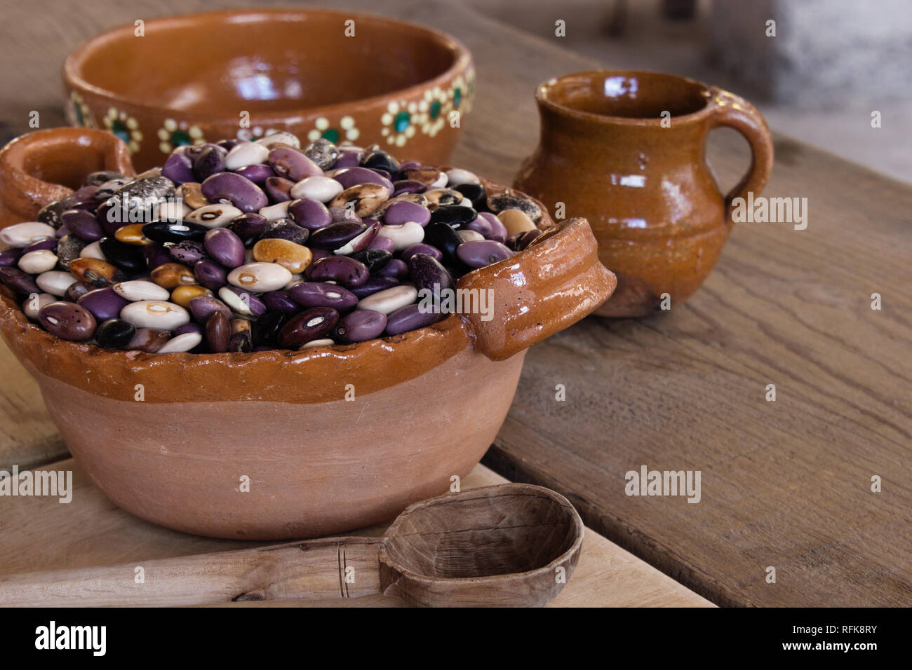 Mexican fatto a mano pentola di creta pieno con un ampia varietà di semi di fagiolo messicano, tazza di argilla, vintage tavola in legno rustico e cucchiaio Foto Stock