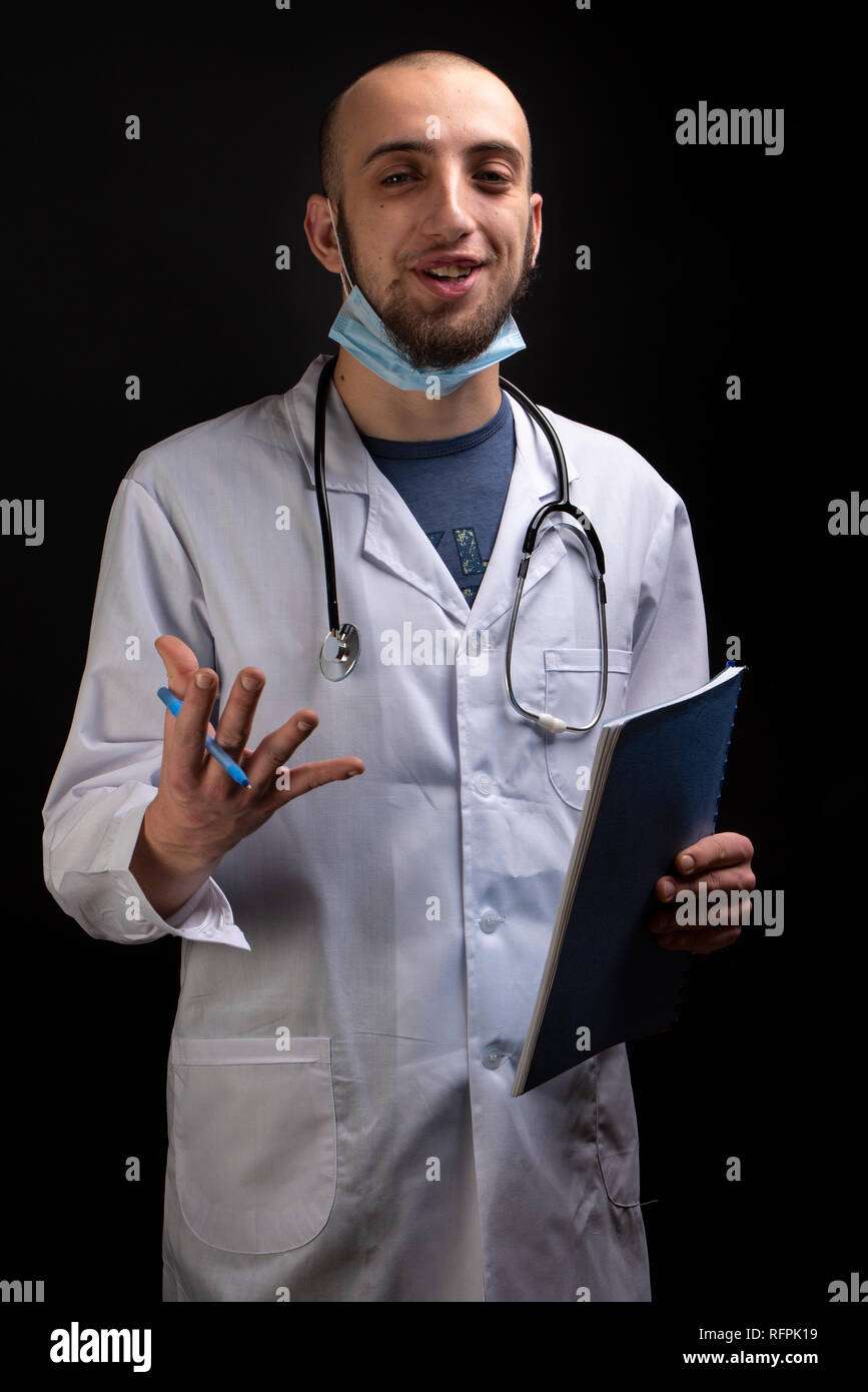 Sorridente giovane medico con stetoscopio, maschera tenendo un file e la visualizzazione di un contenuto di espressione. Specialista di allegro shot su sfondo nero Foto Stock