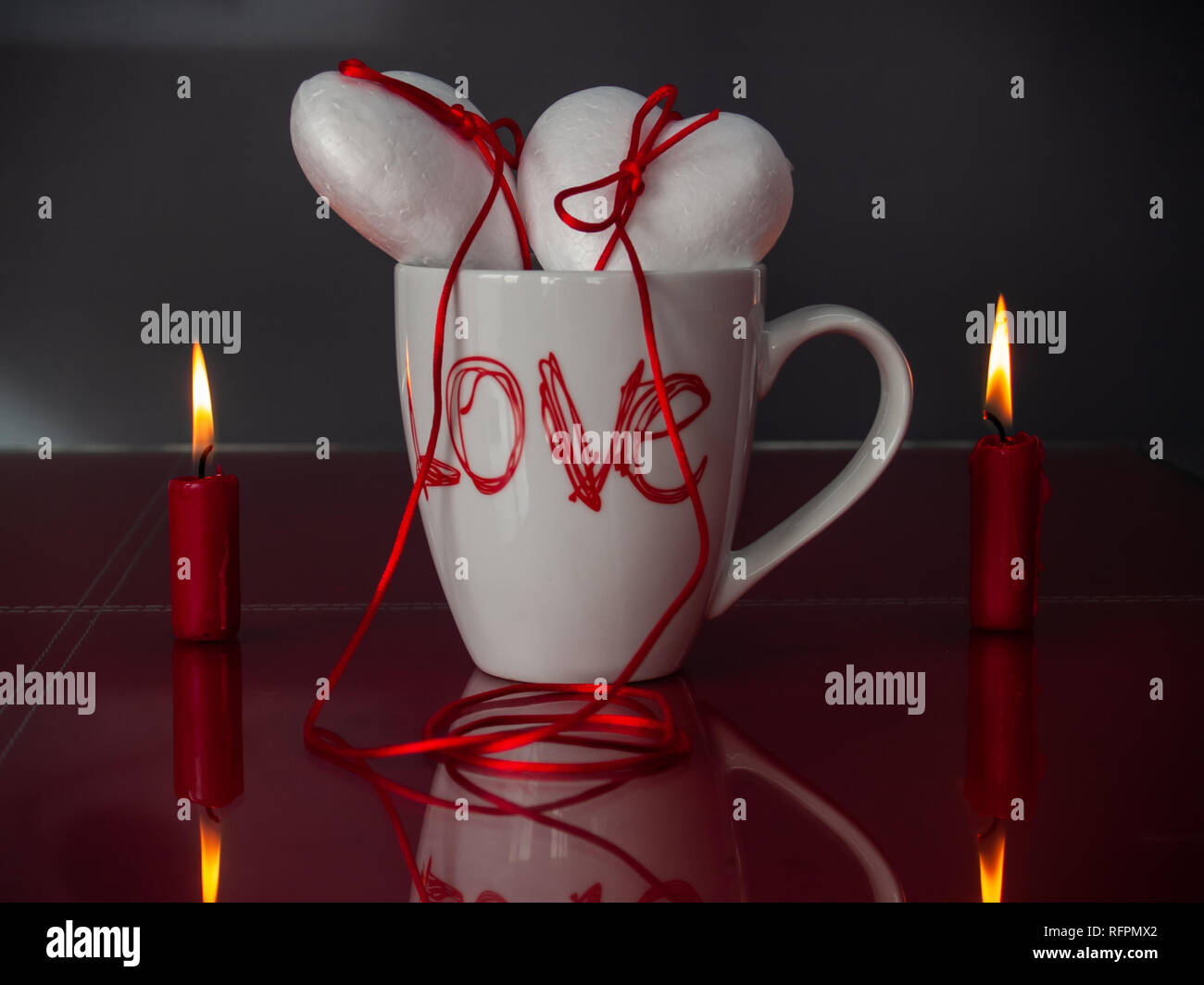 Concetto di amore poliespan due cuori uniti con un filo rosso che simboleggia la leggenda del filo rosso su una tazza colazione e due rosso acceso candele Foto Stock