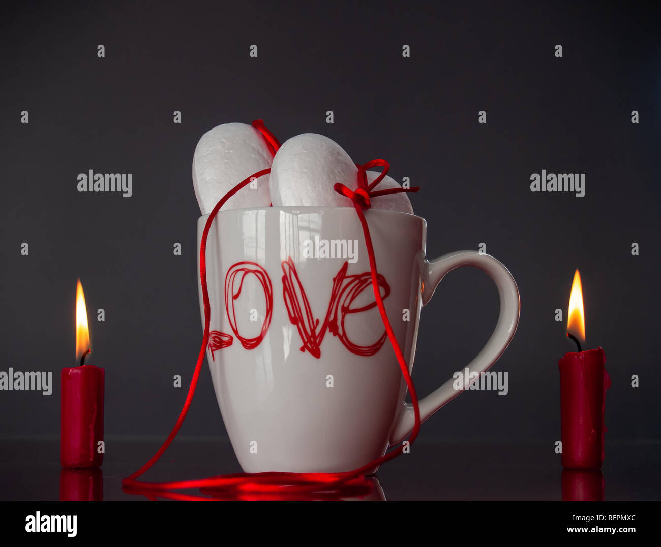 Concetto di amore poliespan due cuori uniti con un filo rosso che simboleggia la leggenda del filo rosso su una tazza colazione e due rosso acceso candele Foto Stock