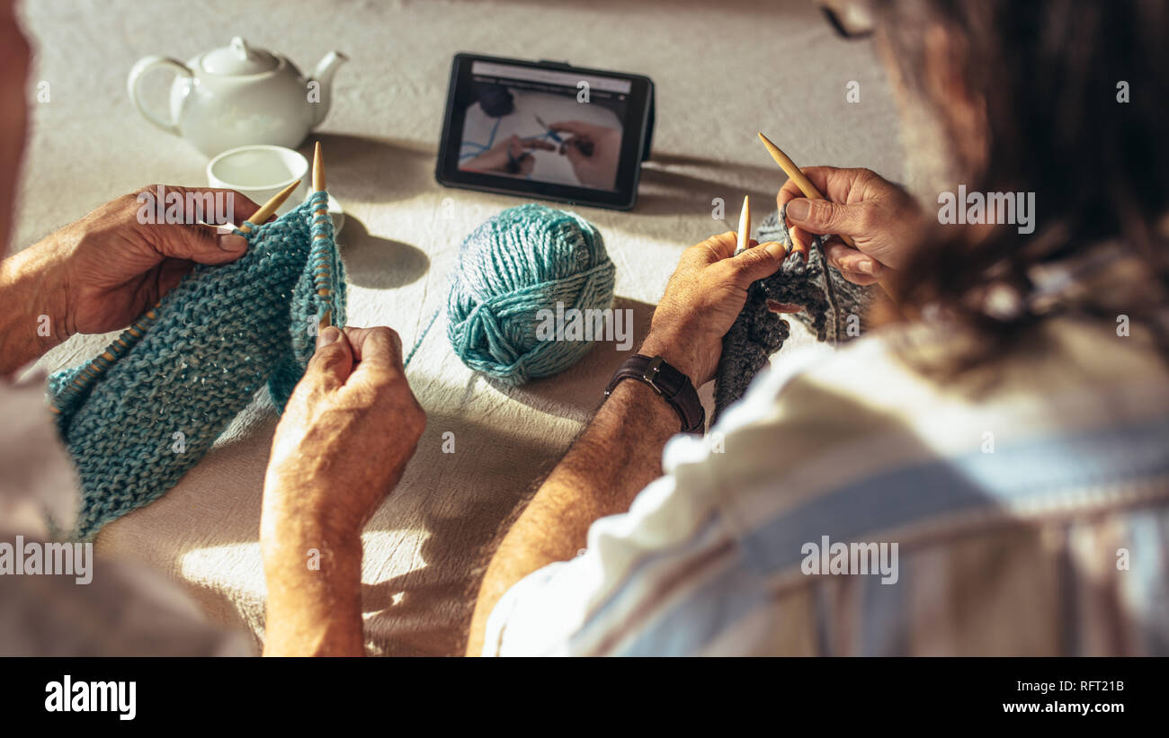 Le mani di alti uomini che tengono degli aghi di tessitura e di lana cantiere seduta a tavola e maglieria di apprendimento dal video online su tavoletta digitale. Pensionati Foto Stock