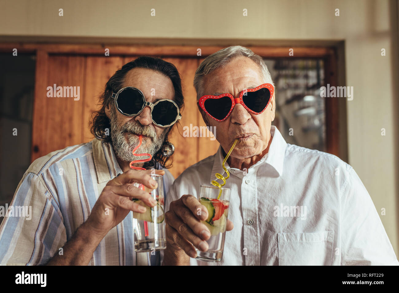 Alti uomini indossando occhiali da sole divertente bere il succo con la paglia. Amici anziani con crazy eyewear avente il succo in ambienti chiusi. Foto Stock