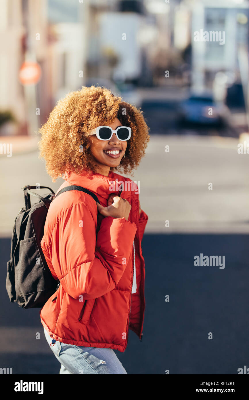 Donna afro american viaggiatore in vacanza in giro per la città. Sorridente turista donna che indossa gli occhiali da sole e uno zaino camminando sulla strada. Foto Stock