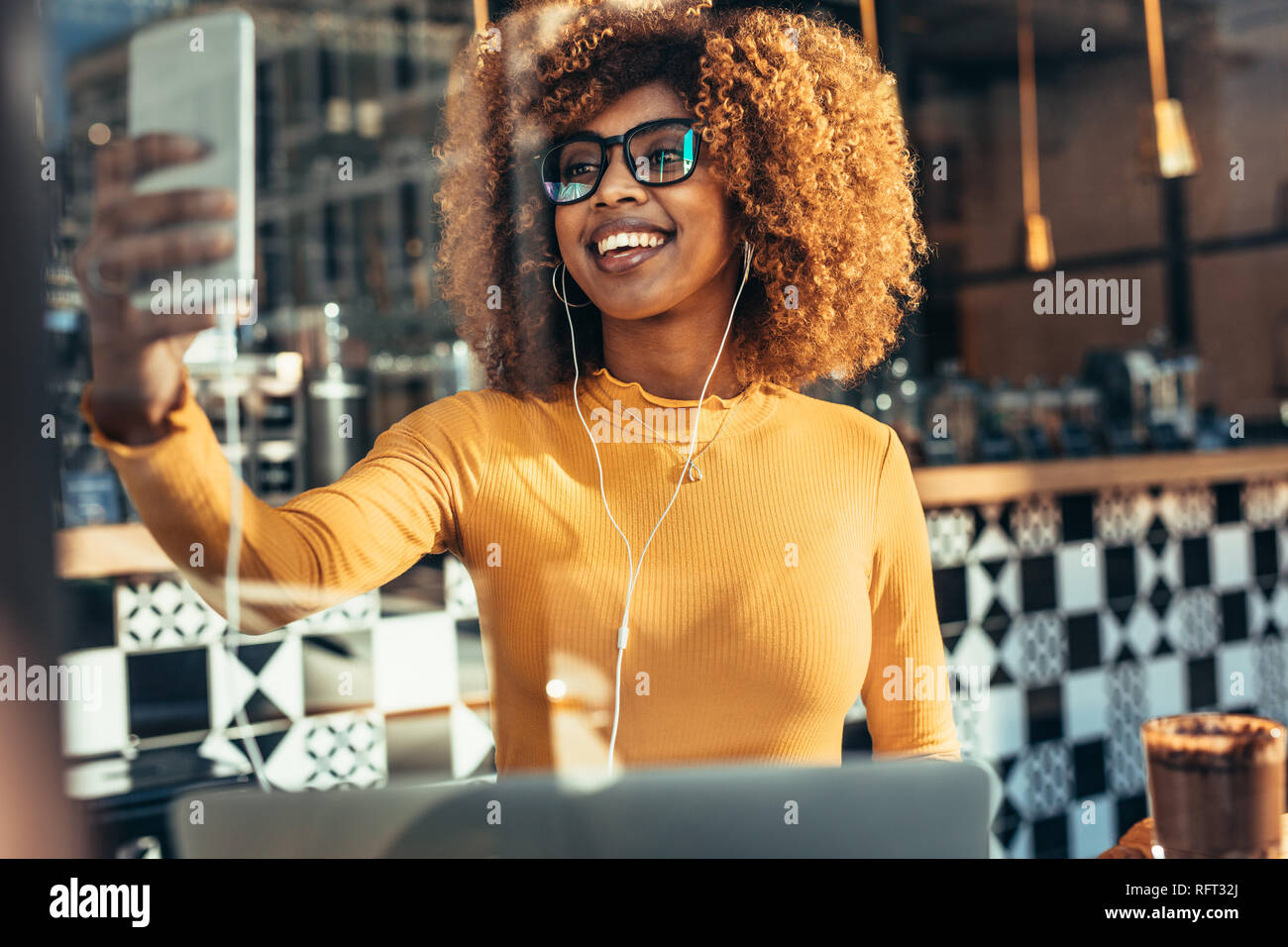 Allegra donna prendendo un selfie tramite telefono cellulare seduti ad un coffee shop. Ritratto di una donna sorridente guardando il suo telefono cellulare mentre ascolti t Foto Stock
