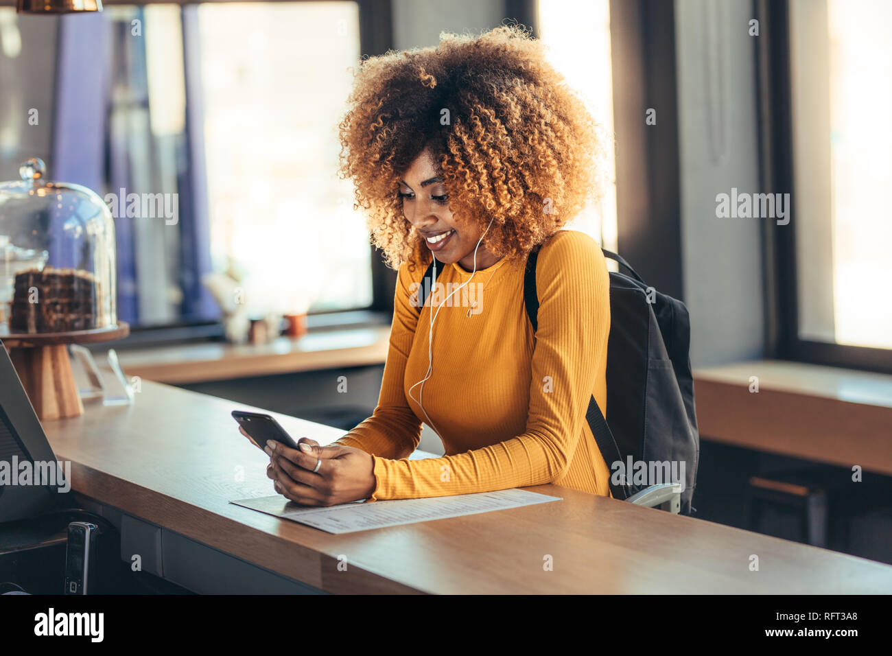Allegro afro american femmina zaino da indossare in piedi al contatore di fatturazione guardando il suo telefono cellulare. La donna in attesa presso il contatore a pagamento di un Foto Stock