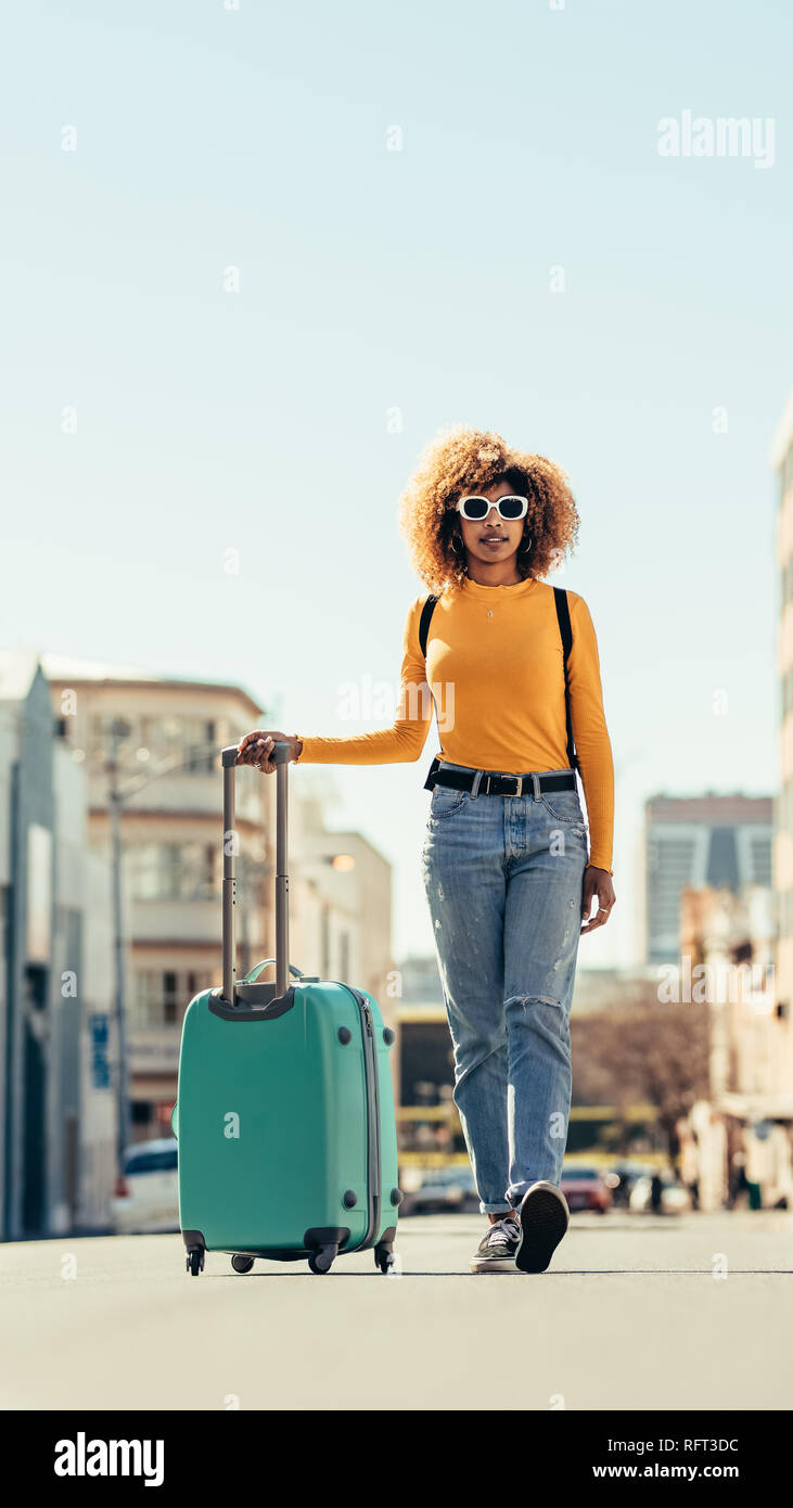 Afro American donna viaggiatore a piedi su strada tenendo un carello. Donna in occhiali da sole che porta uno zaino e TROLLEY BAG a piedi attorno al Foto Stock