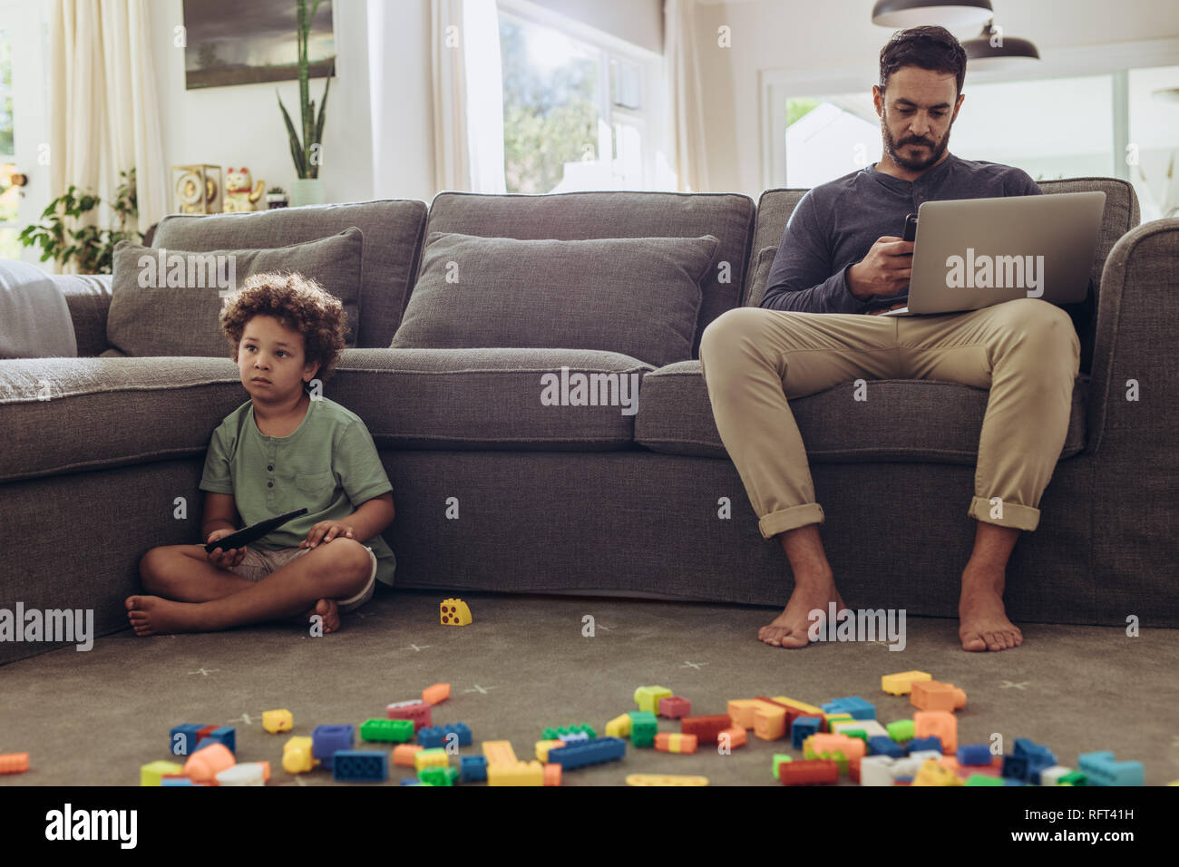 Uomo al lavoro su laptop da casa seduti sul divano. Ragazzo seduto sul pavimento con la costruzione di blocchi davanti e suo padre lavora sul computer portatile. Foto Stock