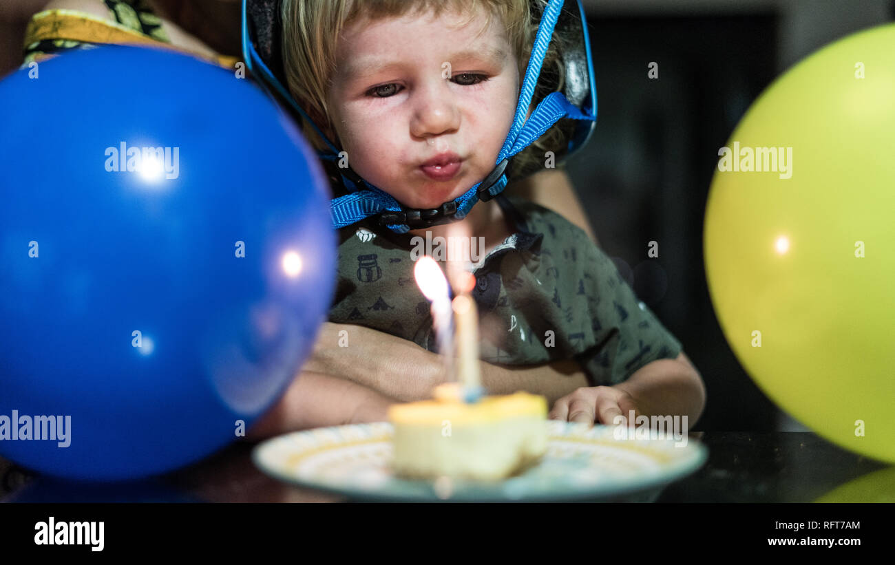 Un ritratto di un felice compleanno ragazzo che indossa un casco blu e soffiando fuori le candeline di compleanno. Blu e gialla aria palloncini in primo piano. Foto Stock