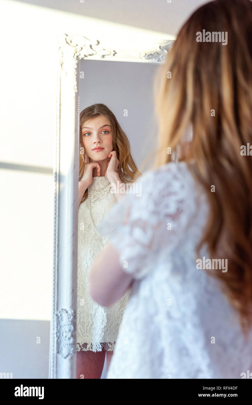 Bellezza, igiene, mattina e concetto di persone - felice sorridente ragazza adolescente guardando riflesso in uno specchio. positivo giovane donna che indossa abito bianco pos Foto Stock