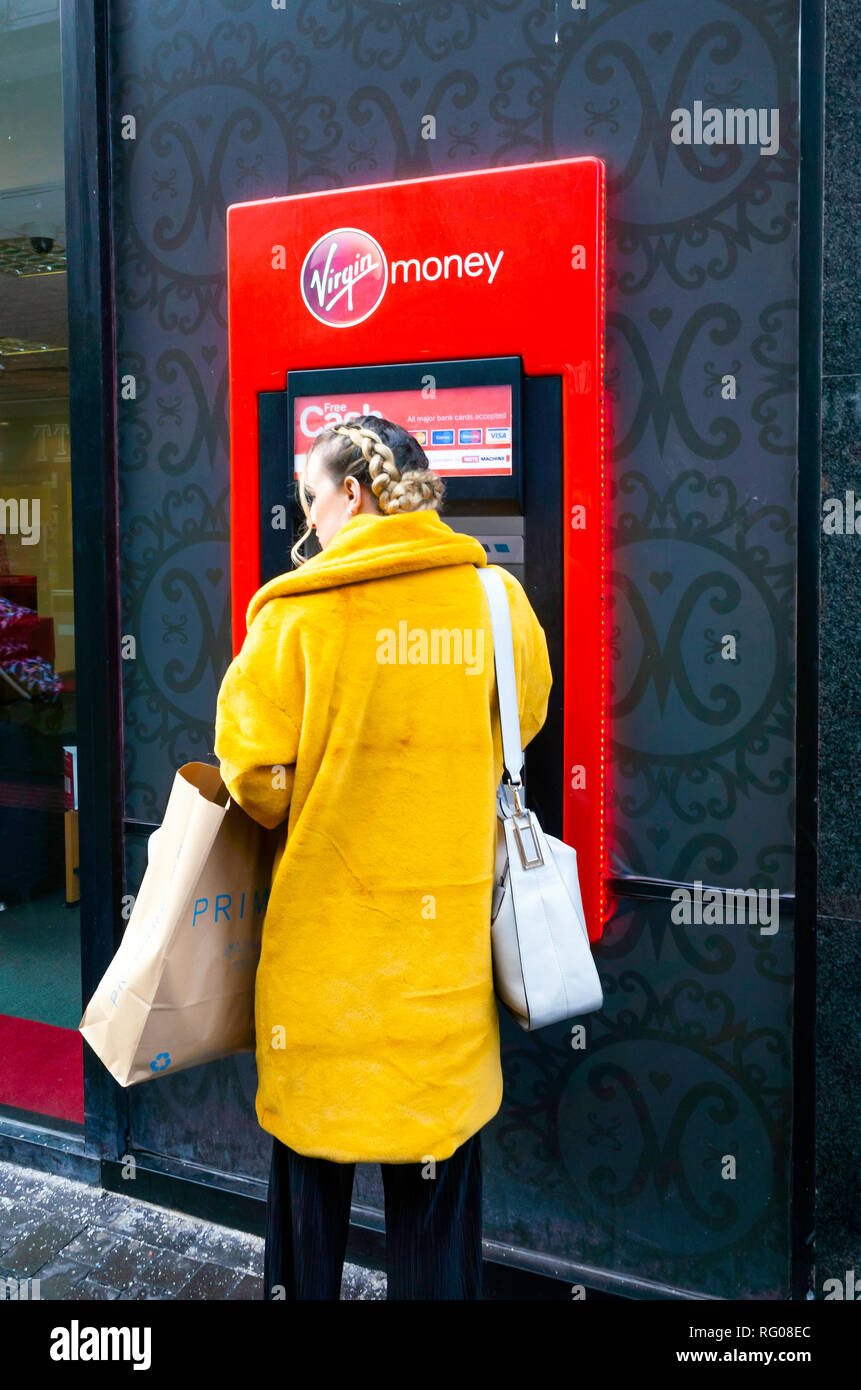 Giovane donna in un rivestimento di colore giallo con una borsa a mano e shopping a una vergine denaro ATM Bancomat Foto Stock