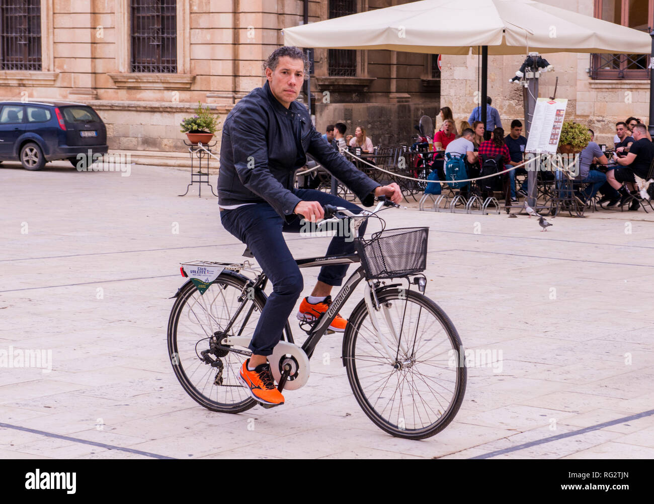 Ritratto di uomo in bicicletta Ciclismo su strada, gruppo di persone al di fuori seduta cafe in background, Ortigia, Siracusa, Sicilia, Italia, Europa Foto Stock