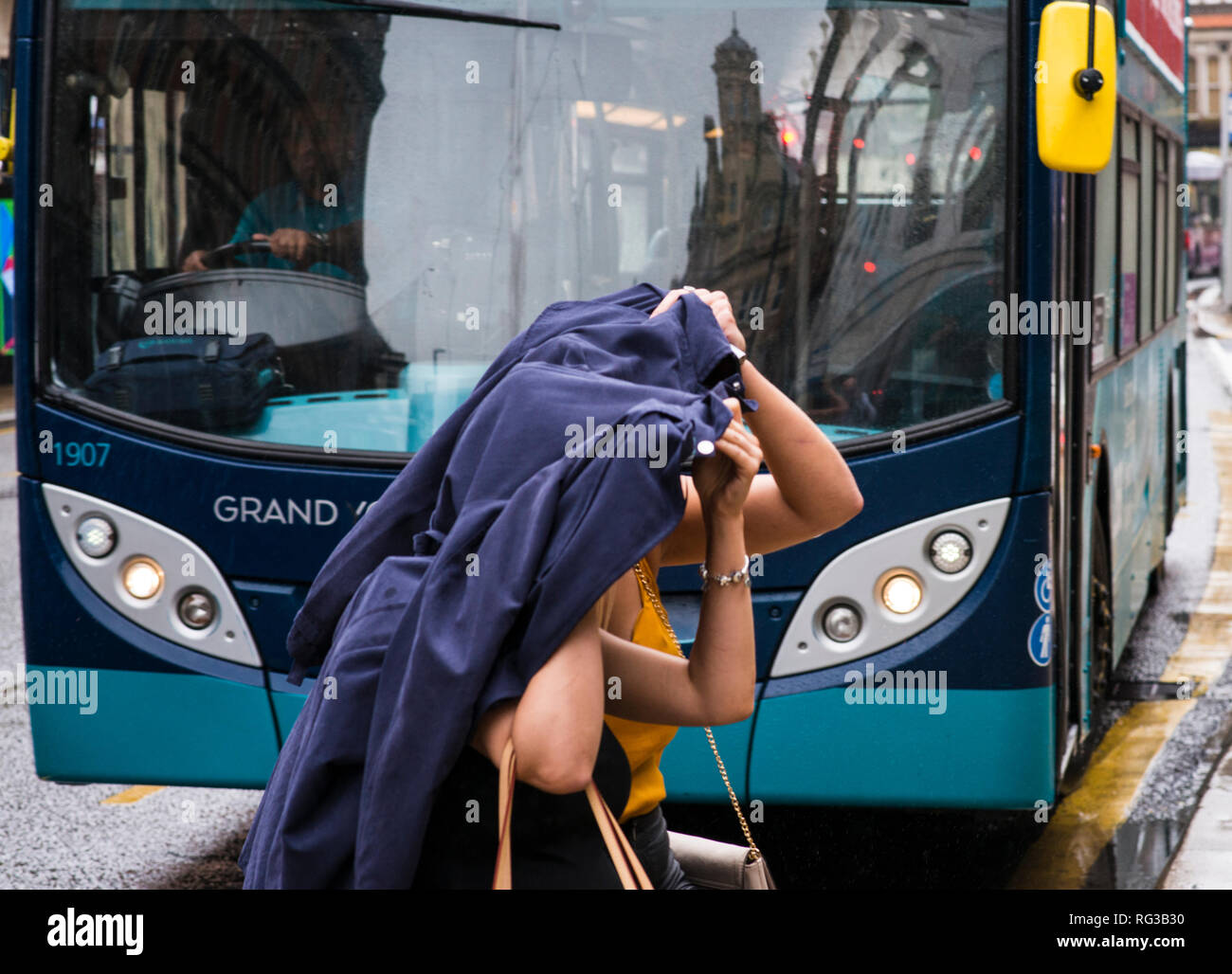 Due donne, attraversamento strada nella pioggia, nella parte anteriore del bus che sopraggiungono, teste di copertura con la camicia, centro di Leeds, nello Yorkshire, Inghilterra, Regno Unito Foto Stock