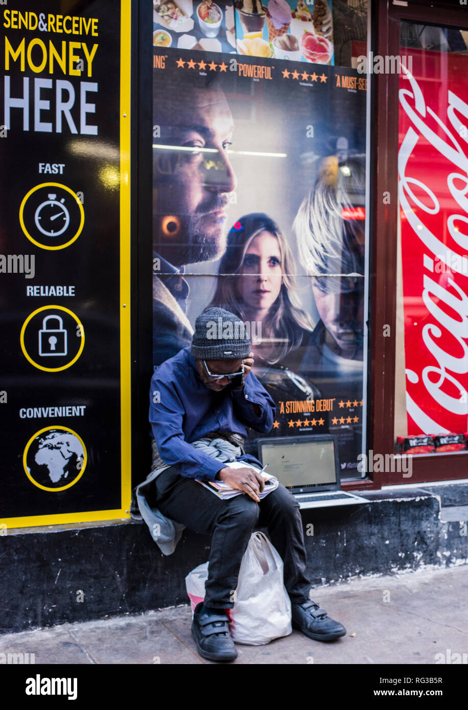 Uomo seduto sul muro, manifesti pubblicitari nella finestra dietro di lui, utilizza lo smartphone, laptop sulla parete accanto a lui, Central London, England, Regno Unito Foto Stock