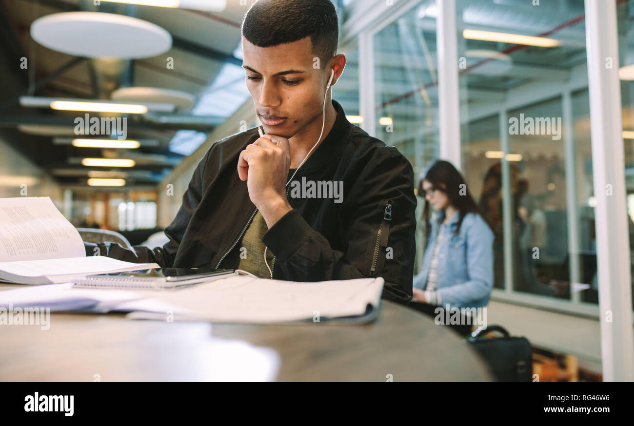 Uomo che indossa gli auricolari guardando a tavoletta digitale mentre è seduto alla biblioteca universitaria. Maschio di studente al college campus utilizzando digitale compressa. Foto Stock