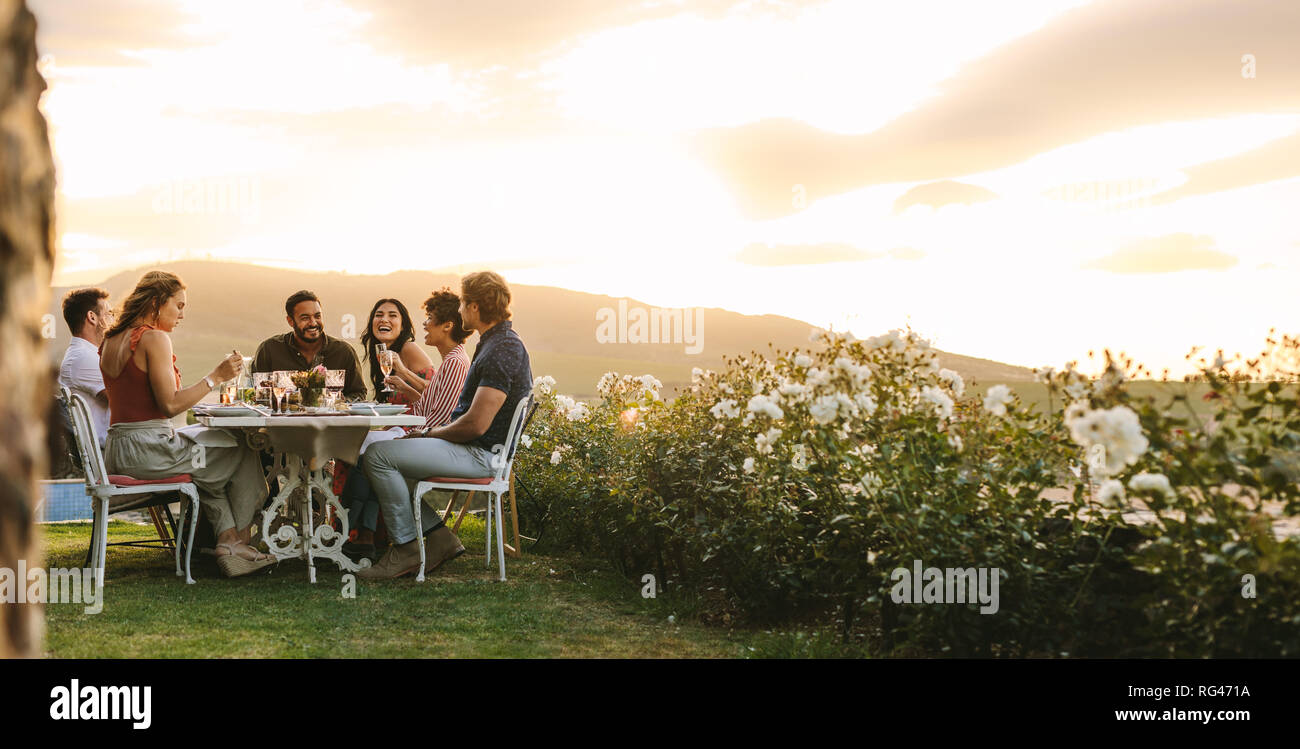 Un gruppo di giovani amici appendere fuori con un drink all'aperto a cena. Giovani uomini e donne seduti attorno a un tavolo di cibo e bevande. Foto Stock