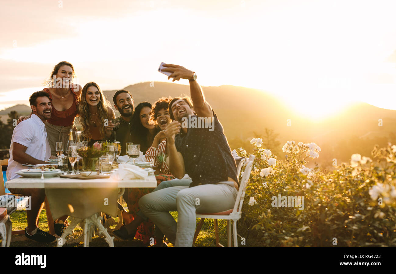 Amici al di fuori di refrigerazione tenendo gruppo selfie e sorridente. Ridere i giovani seduti attorno a un tavolo da pranzo all'aperto e prendendo selfie. Foto Stock