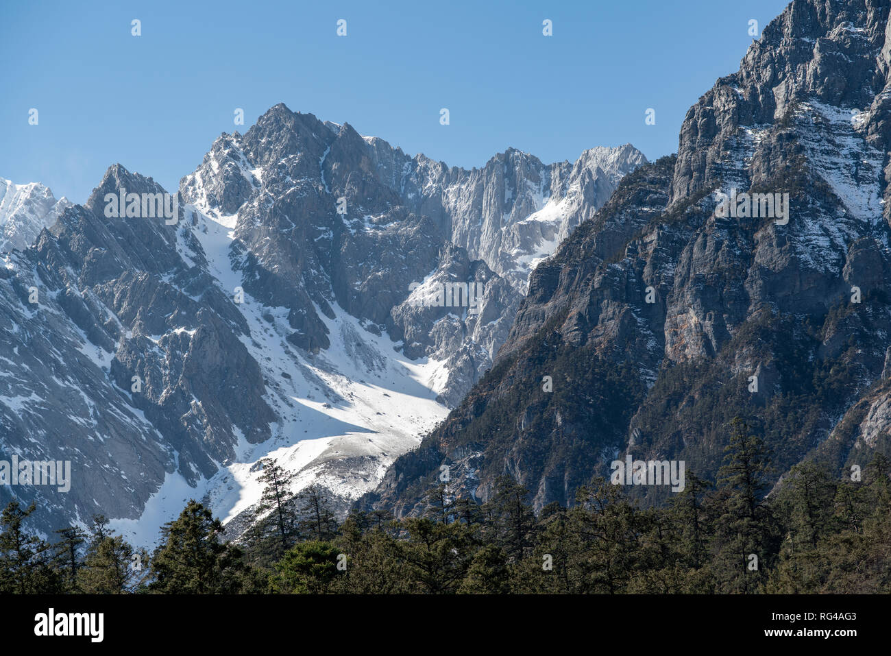 Jade dragon snow mountain situato in Yulong, Yunnan in Cina. Montagne coperte di neve con picchi rocciosi con alberi in primo piano dando una drammatica Foto Stock