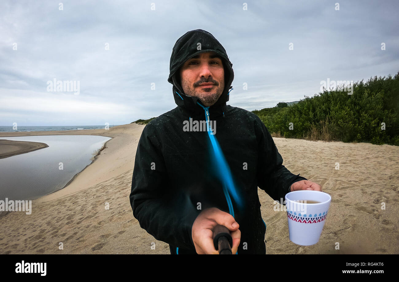 Uomo in piedi sulla spiaggia sabbiosa con selfie stick Sfondo oceano. Ritratto Di Auto dell'uomo in abbigliamento pioggia permanente sulla Atlantic Beach in un giorno di pioggia in wet Foto Stock