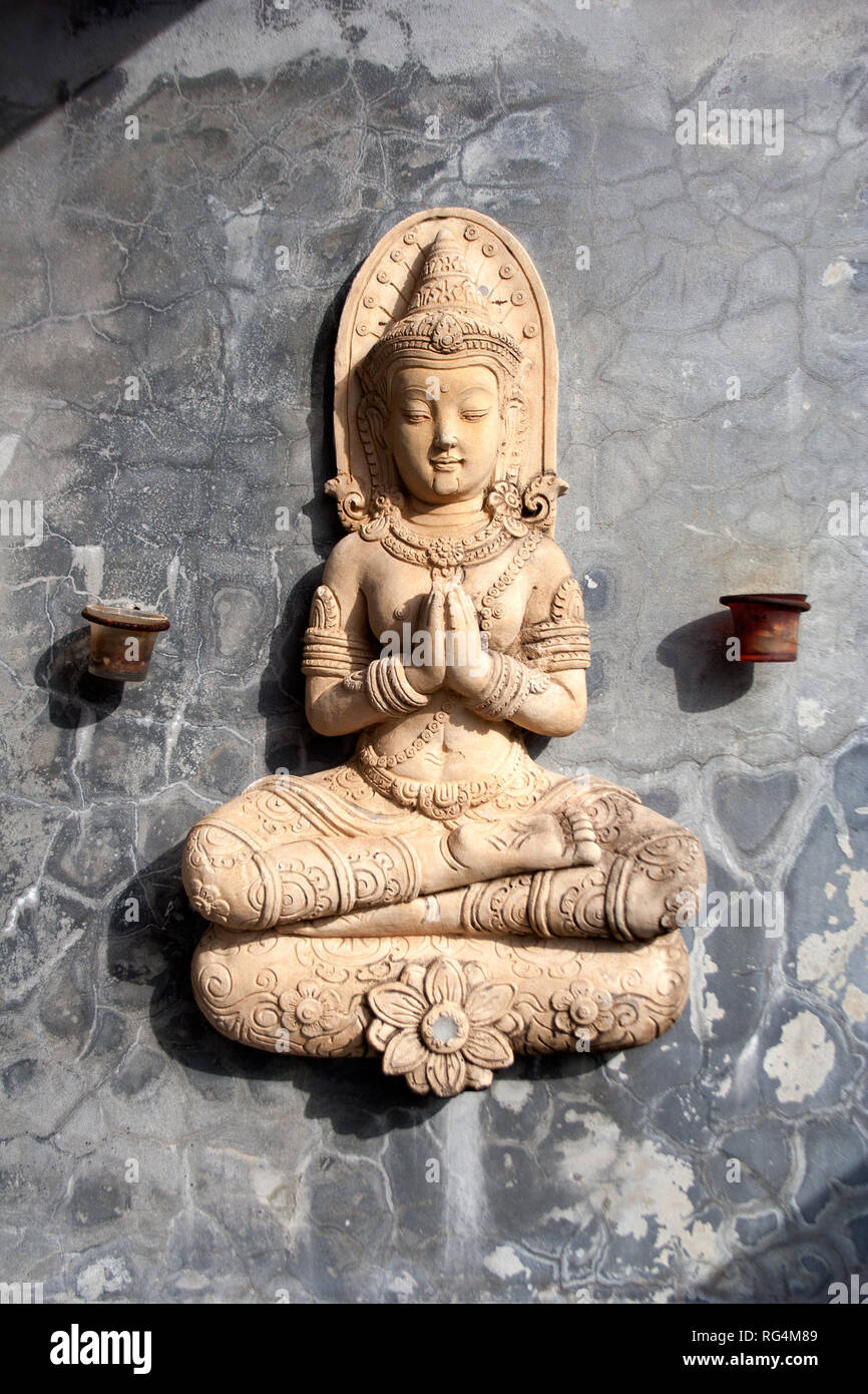 Seduta pregando statua del Buddha, come tradizionale decor tailandese con significato religioso, sul buio muro di pietra con piccoli candelieri sui lati Foto Stock