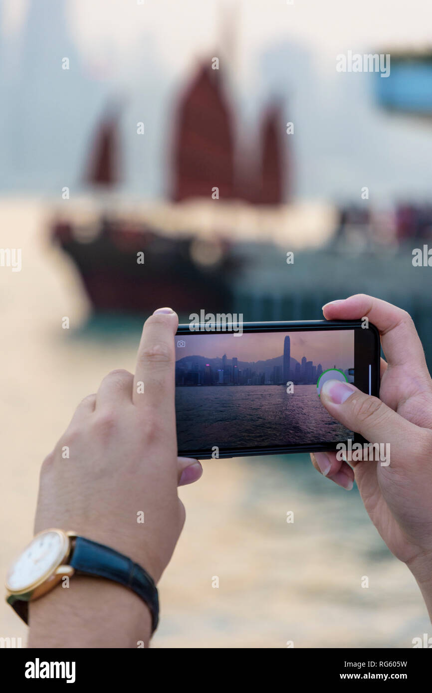 Turistica prendendo una foto sul suo telefono cellulare dello skyline di Hong Kong al tramonto, del porto di Victoria e di Hong Kong Foto Stock