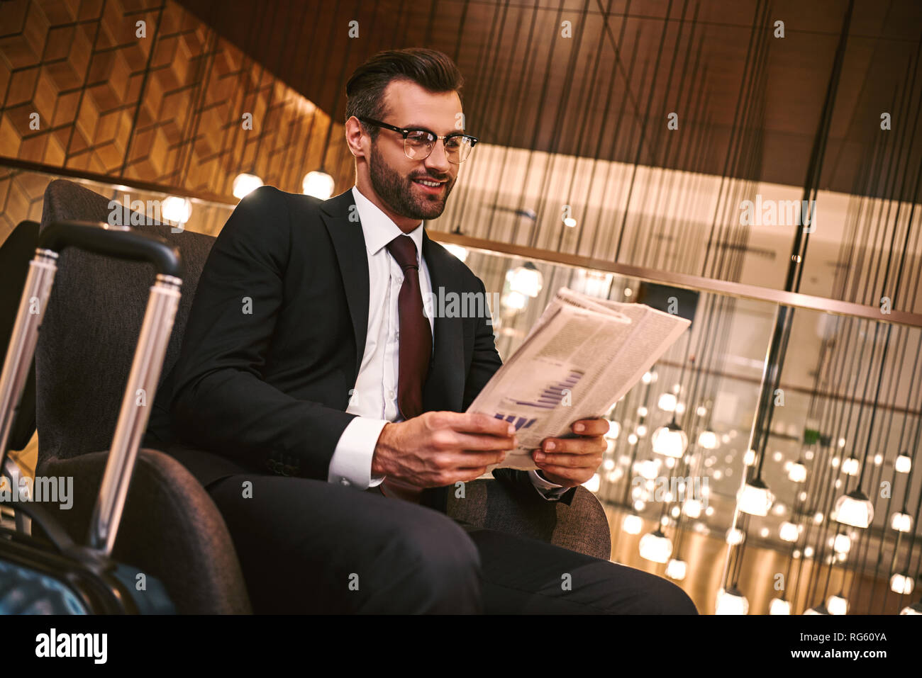In attesa di partner aziendale. Felice giovane uomo che legge il giornale mentre seduti al ristorante dell'hotel vicino a valigia. Lusso a interiour backbround Foto Stock