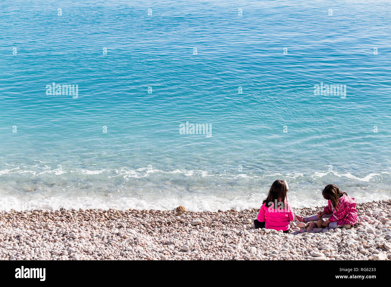 Due ragazze di età compresa tra circa 8 anni seduto su di una spiaggia di ciottoli che indossa giacche rosa con il blu del mare in Spagna. Foto Stock