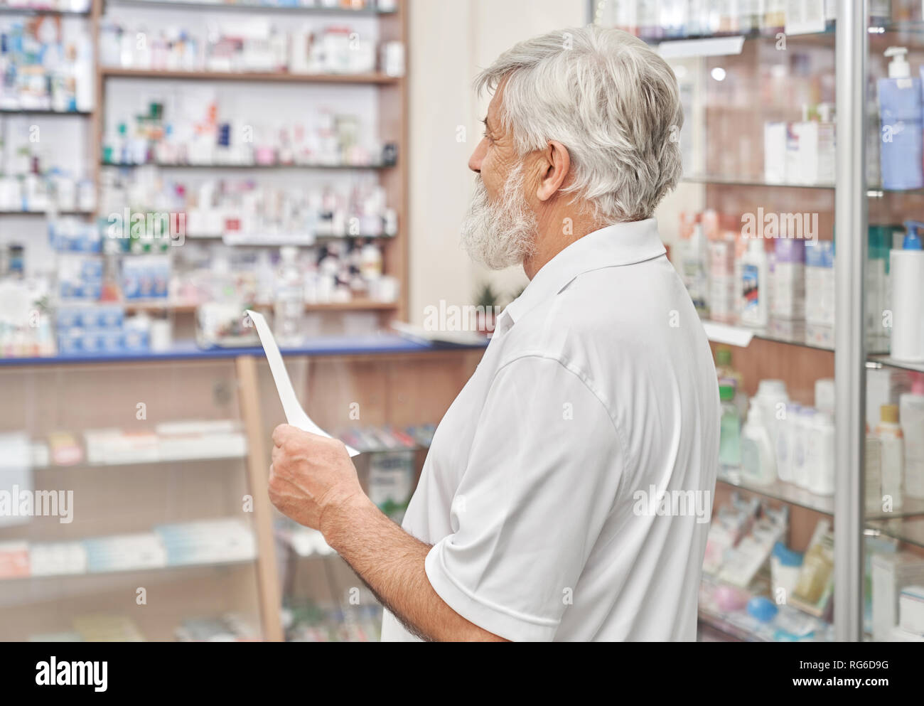 Senior uomo con i capelli grigi e la barba in piedi in farmacia con prescrizione in mano. Il vecchio uomo alla ricerca di farmaci e porre rimedio in farmacia store. Titolare di pensione o di rendita guardando farmacia ripiani. Foto Stock
