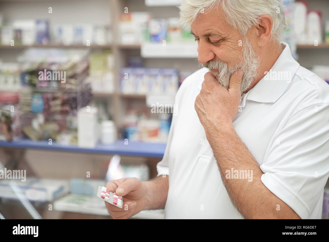 Titolare di pensione o di rendita in piedi in farmacia, tenendo blaster packwith pillole. Uomo anziano pensando, scegliendo i farmaci di malattia. Vecchio cliente acquisto di farmaci per la cura della salute. Foto Stock