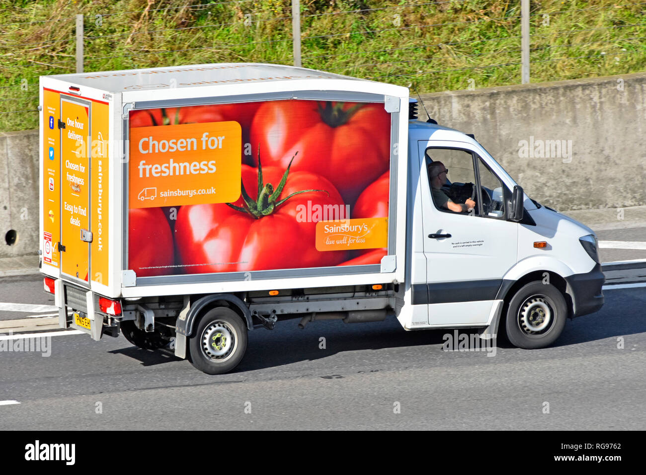 Guardando verso il basso e laterale vista posteriore supermercato Sainsburys home Consegna del cibo van & driver con veicolo pubblicitario di pomodori freschi la guida su autostrada DEL REGNO UNITO Foto Stock