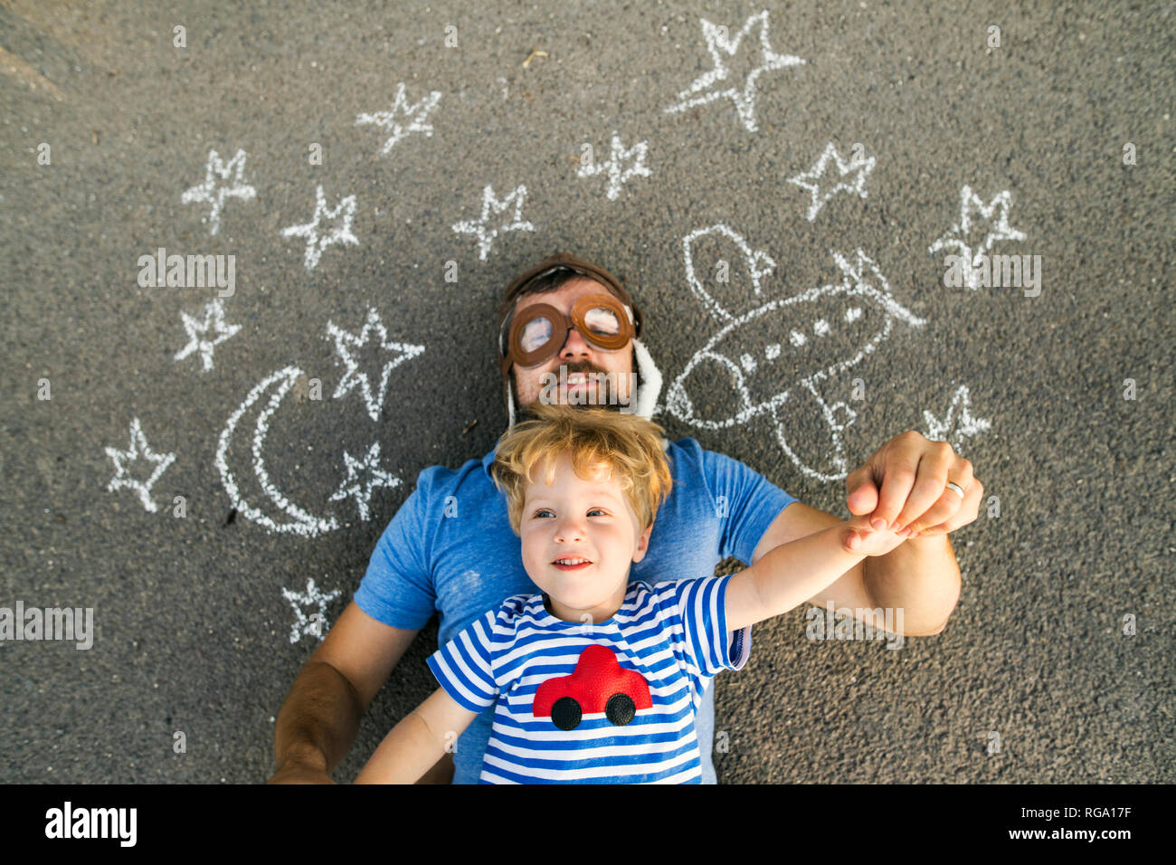 Ritratto di uomo maturo indossando cappello pilota e il suo piccolo figlio giacente su asfalto verniciato con aereo, luna e stelle Foto Stock