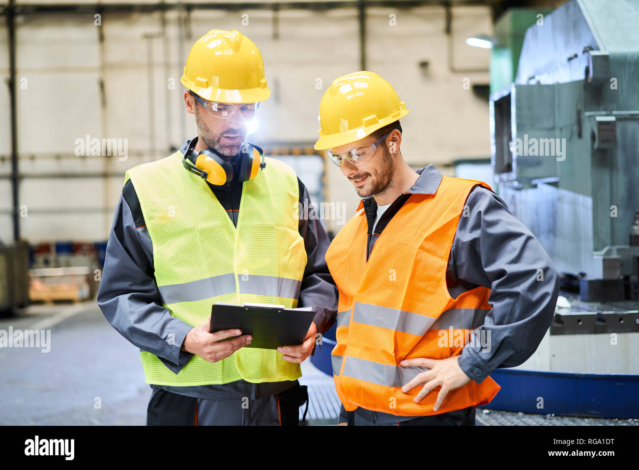 Due uomini che indossano indumenti da lavoro protettiva holding negli appunti e parlando in fabbrica Foto Stock