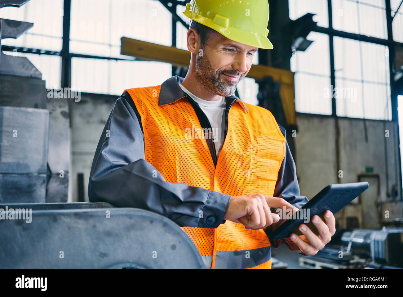 Uomo sorridente indossando indumenti da lavoro protettiva utilizzando tablet in fabbrica Foto Stock