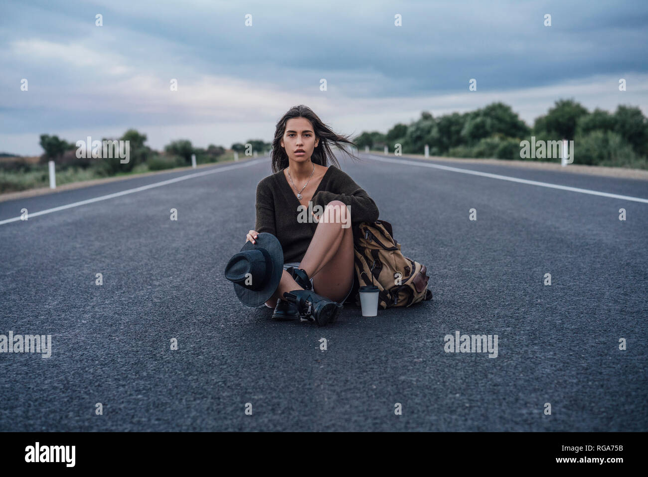 Ritratto di autostop giovane donna con zaino e bevanda seduto su lane Foto Stock