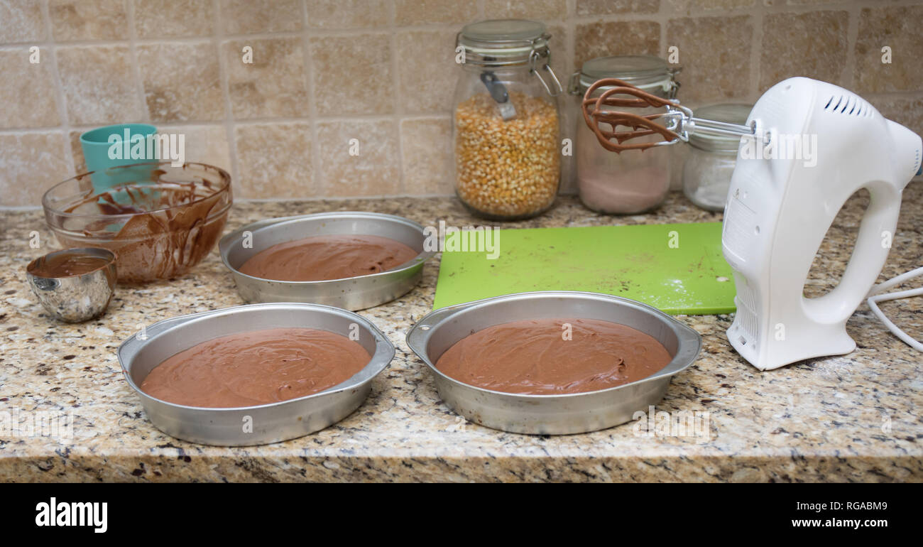 Tre tortiere con torta al cioccolato pastella steso su un banco di cucina con un mixer elettrico. Foto Stock
