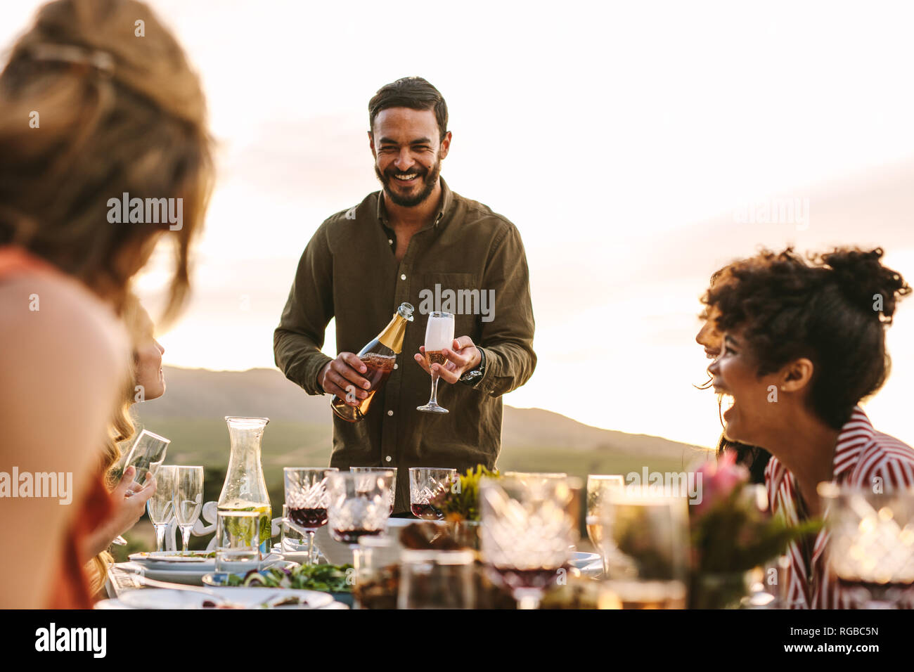 Uomo sorridente che serve champagne per gli amici seduti attorno al tavolo di gioco durante il party. Multi-etnico gruppo di amici avente parte all'aperto. Foto Stock