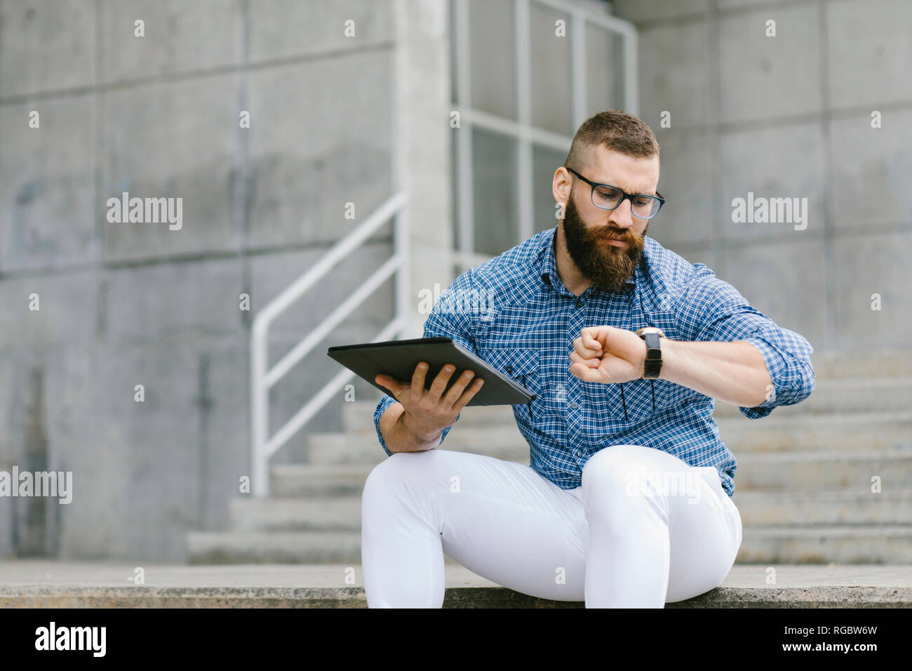 Ritratto di barbuto hipster imprenditore seduti sulle scale con tavoletta digitale controllando il tempo Foto Stock