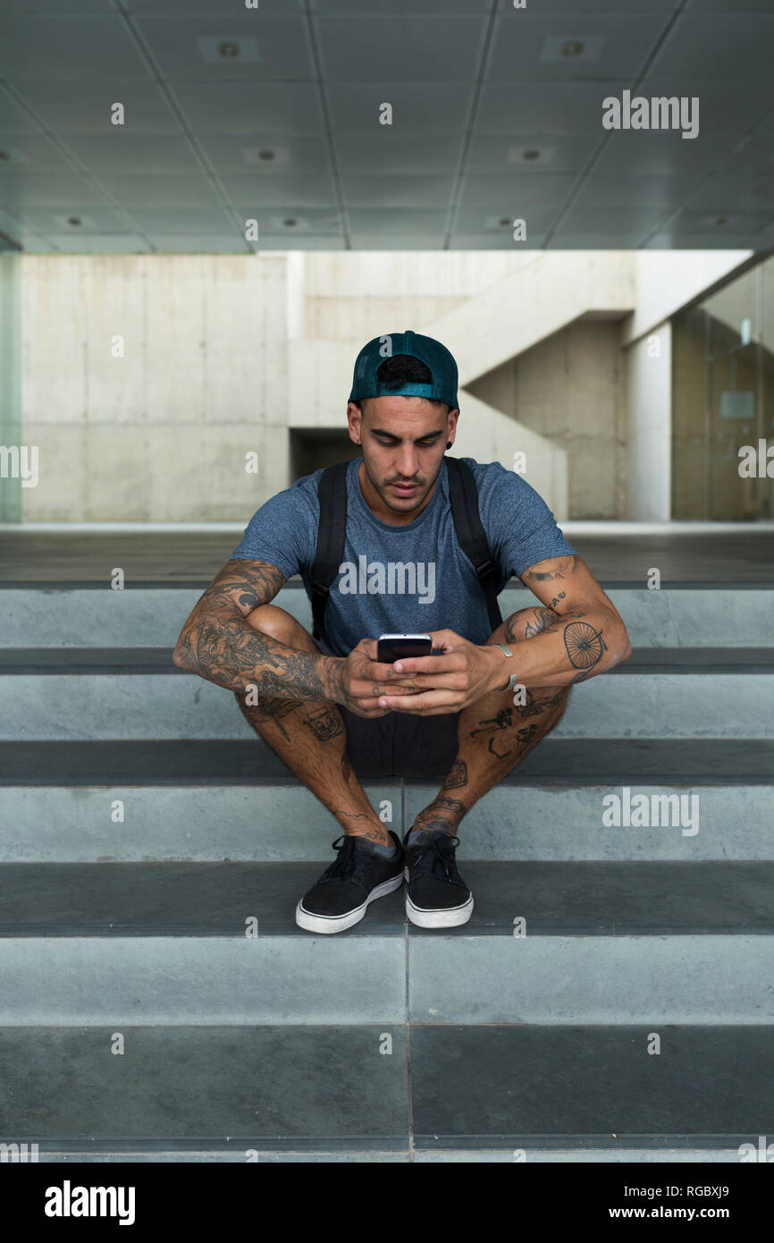 Tatuato giovane uomo seduto sulle scale utilizza lo smartphone Foto Stock