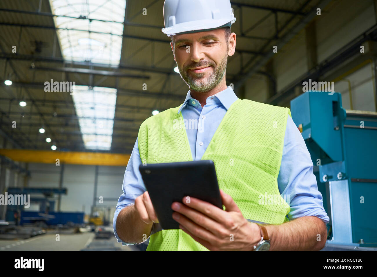 Fiducioso uomo che indossa maglietta e giubbotto di sicurezza utilizzando tablet in fabbrica Foto Stock
