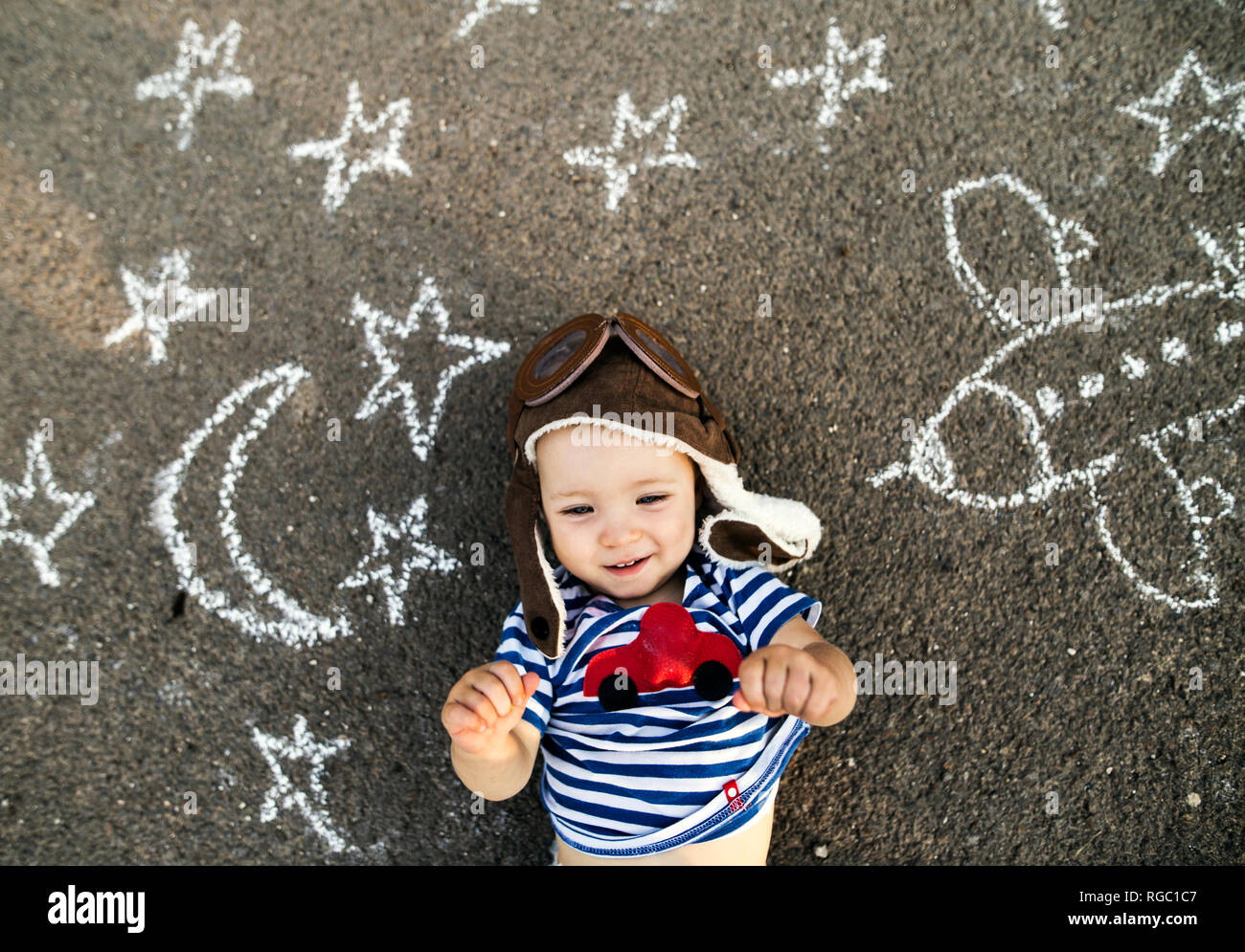 Ritratto di sorridente bambina indossa cappello pilota giacente su asfalto verniciato con aereo, luna e stelle Foto Stock
