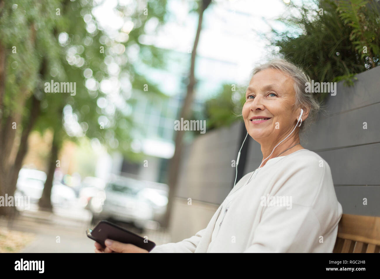 Senior sorridente donna seduta su una panchina all'aperto con un telefono cellulare e auricolari Foto Stock