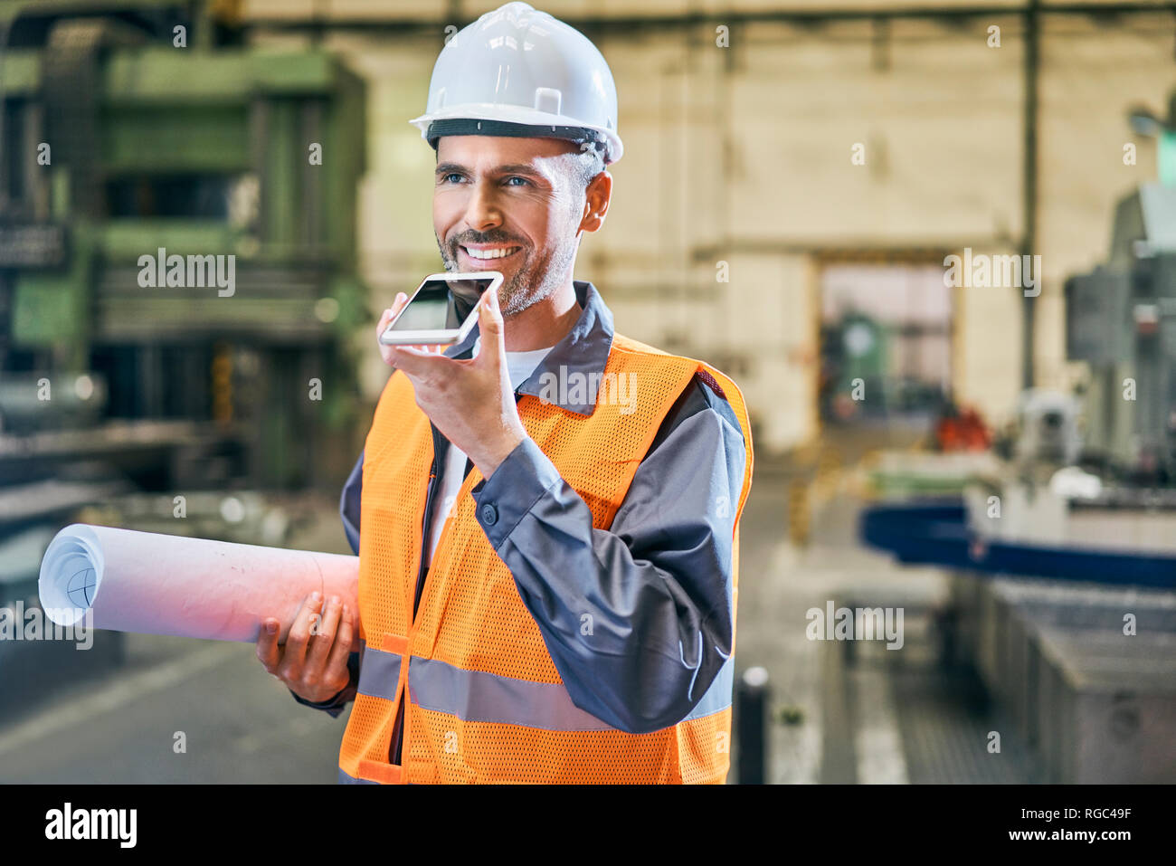 Uomo sorridente holding blueprint e utilizzando il cellulare in fabbrica Foto Stock