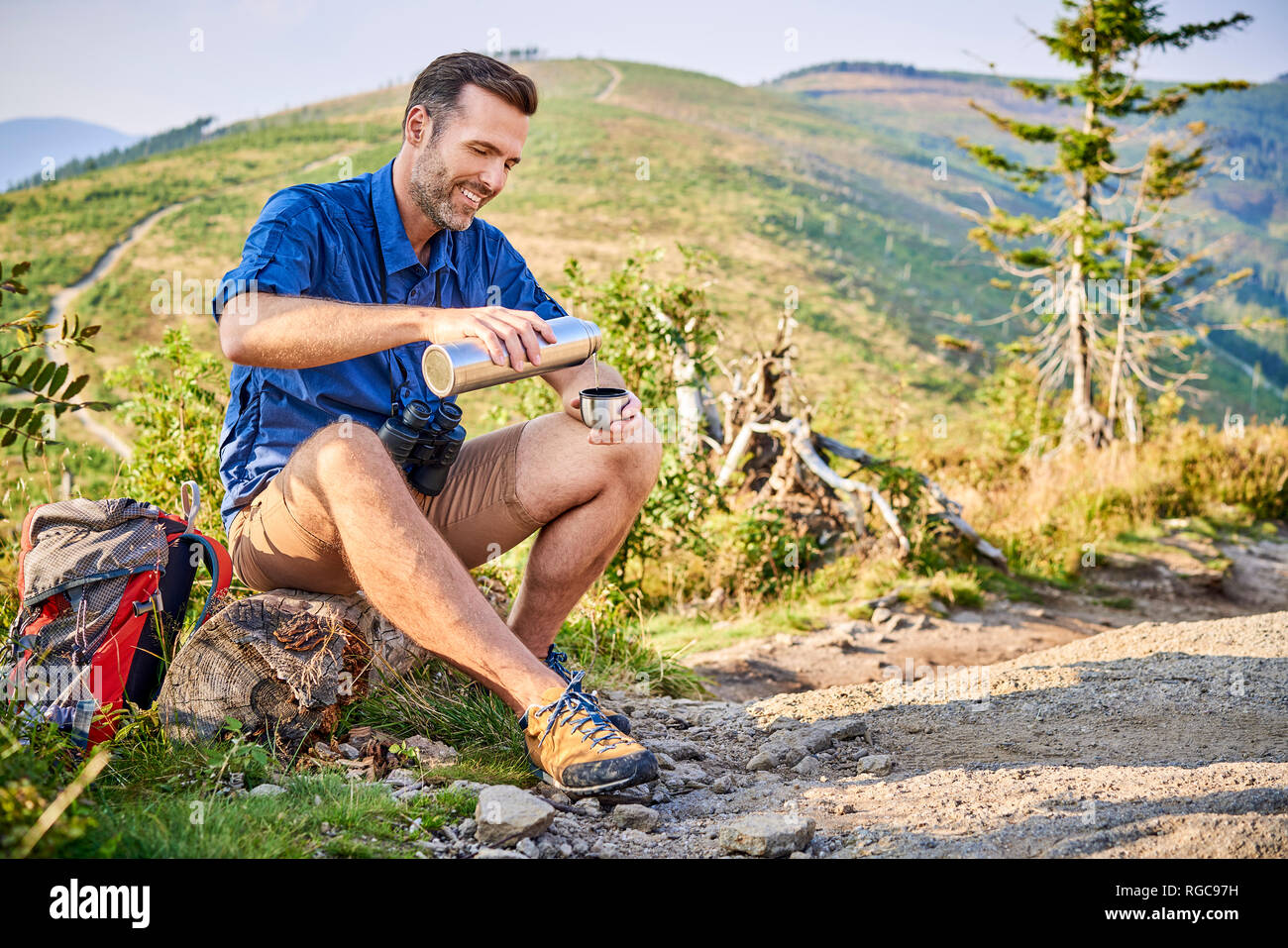 Uomo sorridente in appoggio durante il viaggio escursionistico versando acqua fredda da thermos Foto Stock