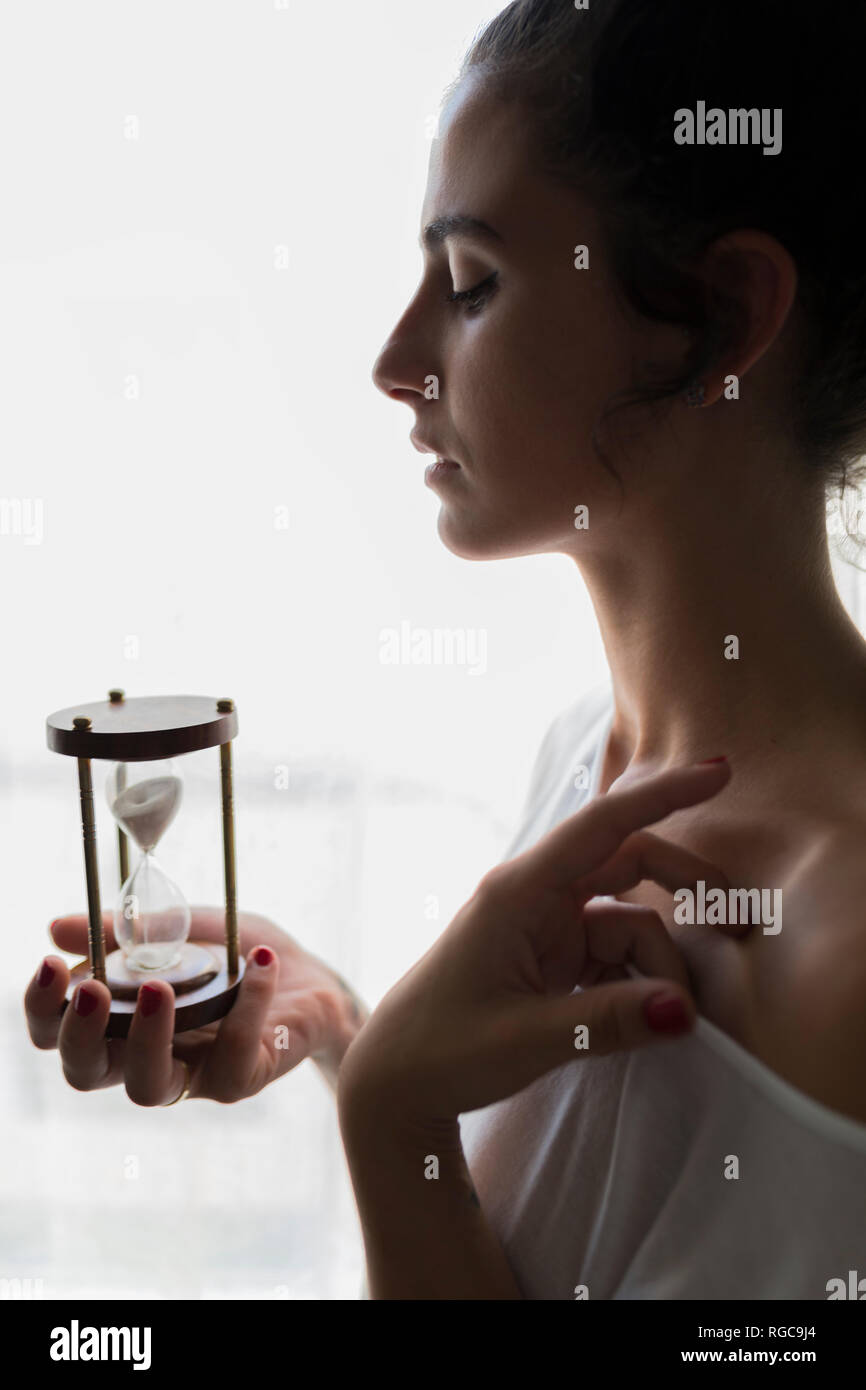 Giovane donna tenendo una clessidra contro una finestra luminosa Foto Stock
