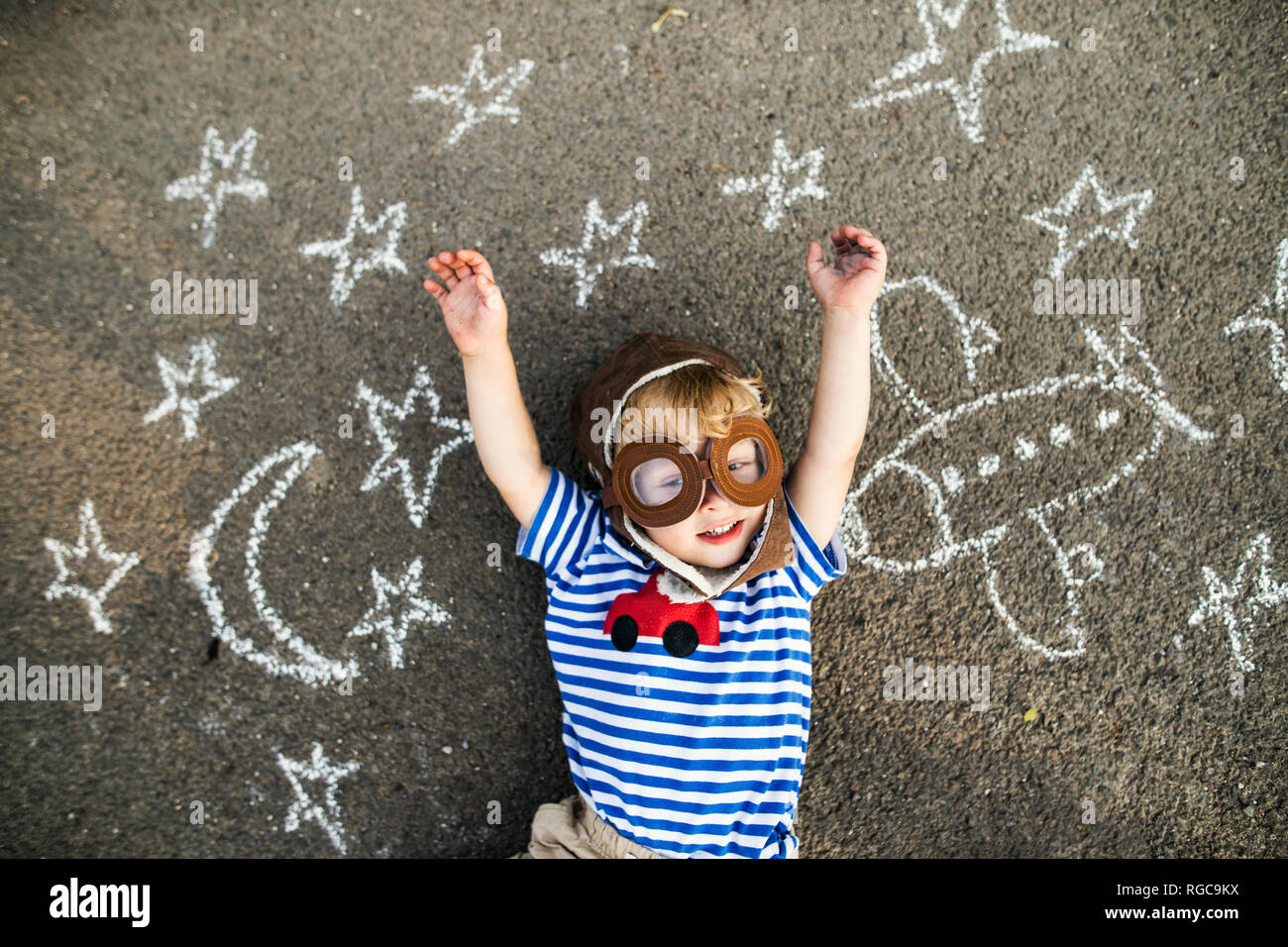 Ritratto di sorriso toddler pilota che indossa un cappello e occhiali giacente su asfalto verniciato con aereo, luna e stelle Foto Stock