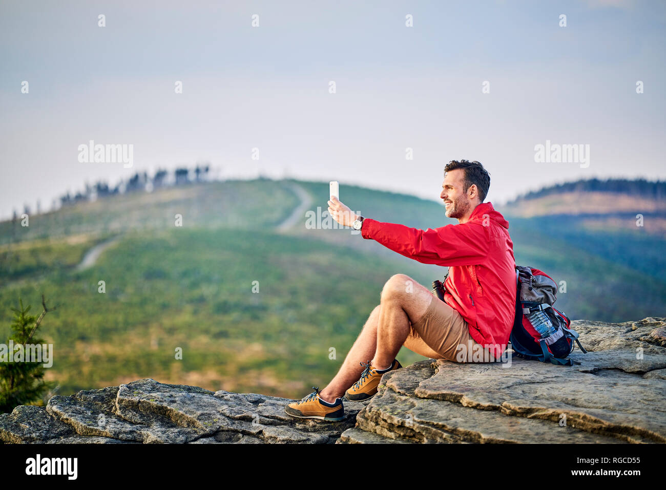 Uomo seduto su roccia tenendo selfie con il suo telefono cellulare durante il viaggio escursioni in montagna Foto Stock