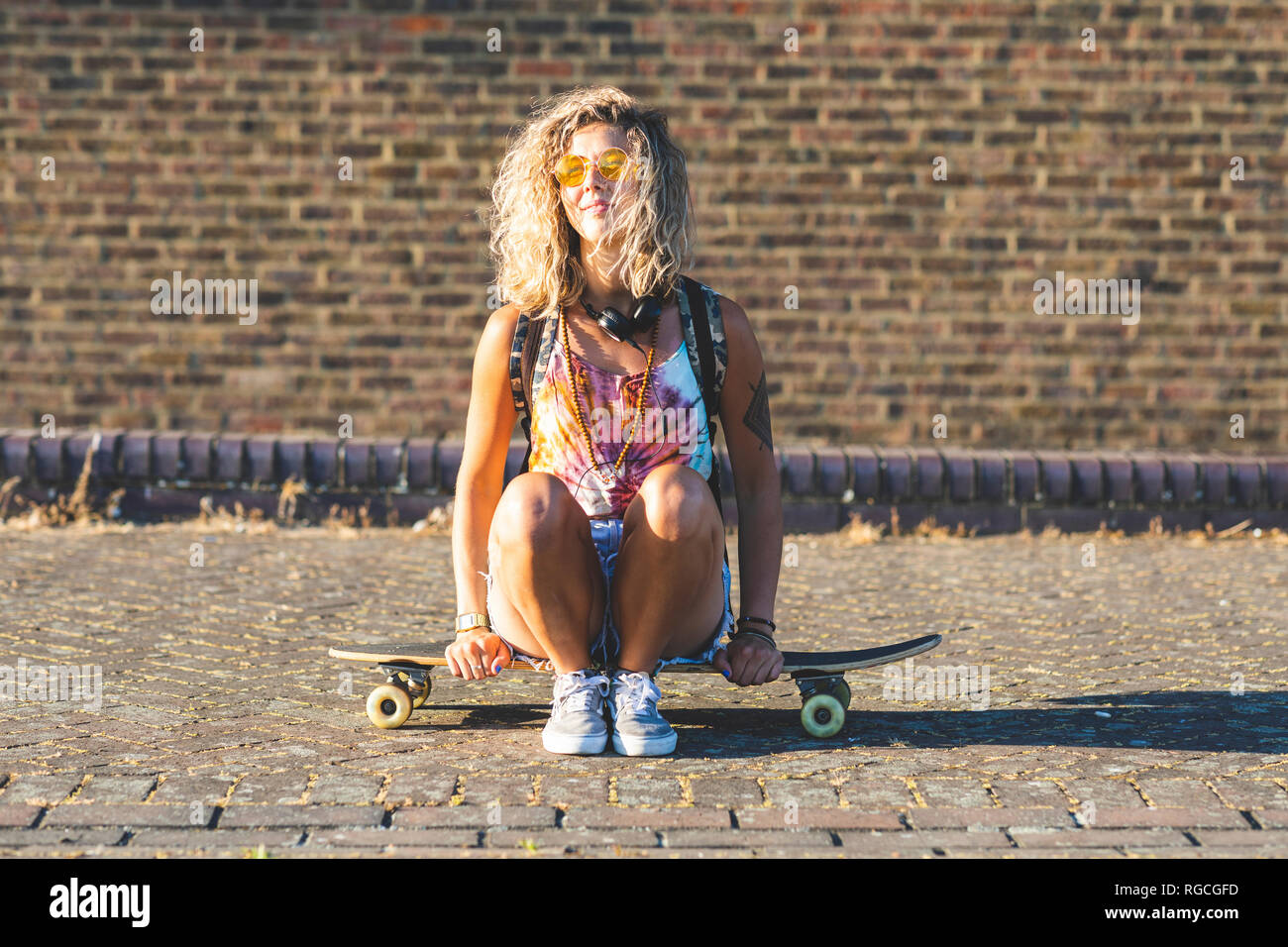 Giovane donna seduta su uno skateboard con un muro di mattoni in background Foto Stock